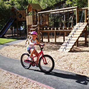 A young girl wearing a pink helmet rides a pink bicycle along a paved path in a playground with wooden climbing structures, slides, and rope bridges surrounded by trees.