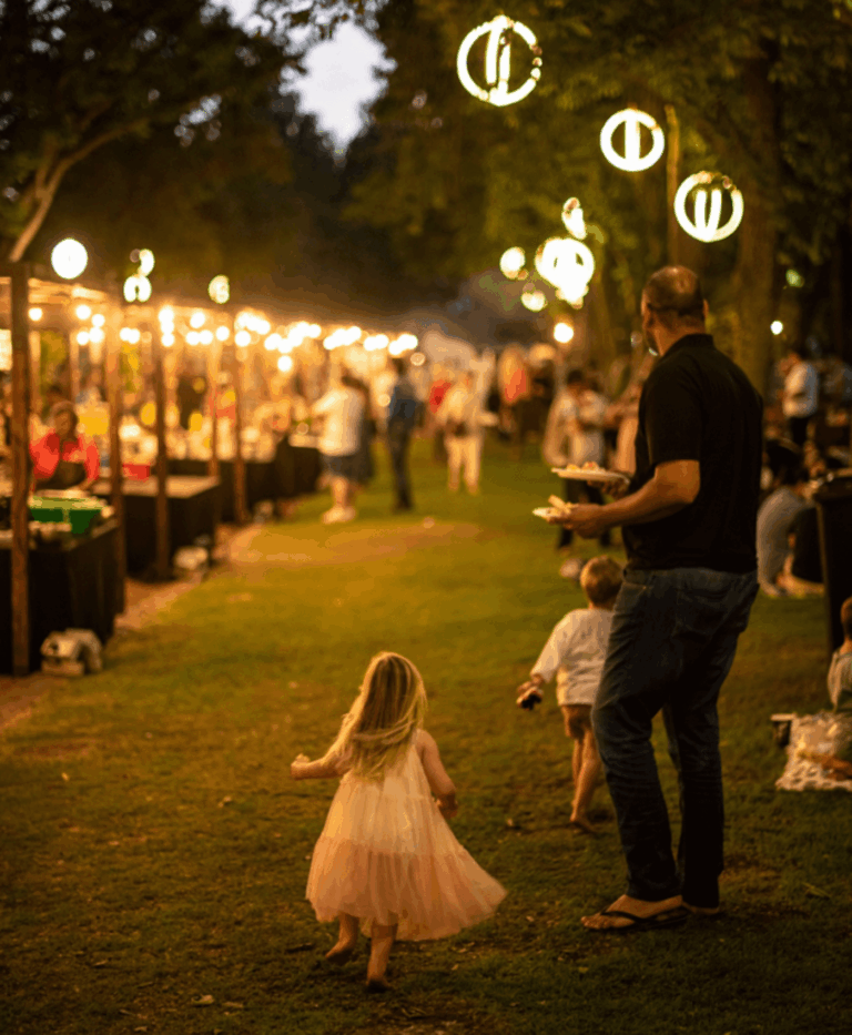 Golden evening light filters through rows of trees strung with glowing orbs, creating a warm and inviting atmosphere. Families wander between stalls lined with twinkling lights as people chat and sample food. A barefoot little girl in a pink and white dress runs ahead on the grass while a man carrying plates follows, capturing a sense of carefree joy at an outdoor night market.