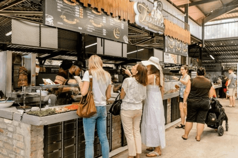 A lively market food stall under a covered structure. A group of people, mostly women, are standing in line at a counter labeled “Tunisian Delights.” The stall displays a vertical rotisserie with meat (likely shawarma) and a colorful spread of fresh toppings. The atmosphere is relaxed and social, with natural lighting and an open layout, typical of a weekend market or food hall.