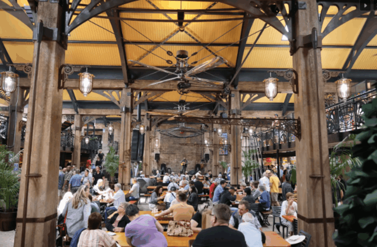 A bustling indoor market or food hall with a warm, industrial aesthetic. The space features tall wooden pillars, metal trusses, and a yellow ceiling that gives the scene a cozy glow. People are gathered at communal wooden tables, chatting and enjoying food and drinks. The layout includes an elevated balcony level, leafy plants adding greenery, and a musician performing live on stage in the background, creating a lively, social atmosphere.