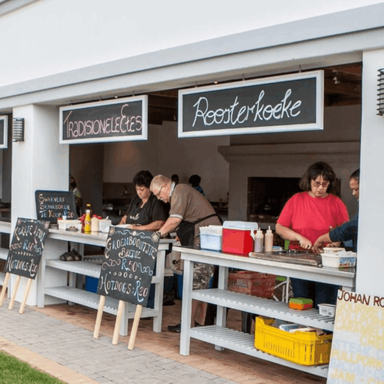 This image shows a vibrant local food stall setup serving traditional South African dishes. Three people are preparing food behind counters labeled **“Tradisionele Etes”** (Traditional Meals) and **“Roosterkoeke”** (grilled bread rolls). Handwritten chalkboard signs advertise items like *Groenboontjie bredie* (green bean stew), *hotdogs*, and *swarwer skinkborde*. The setup has a welcoming, community market atmosphere, with simple wooden tables and bright, casual energy.