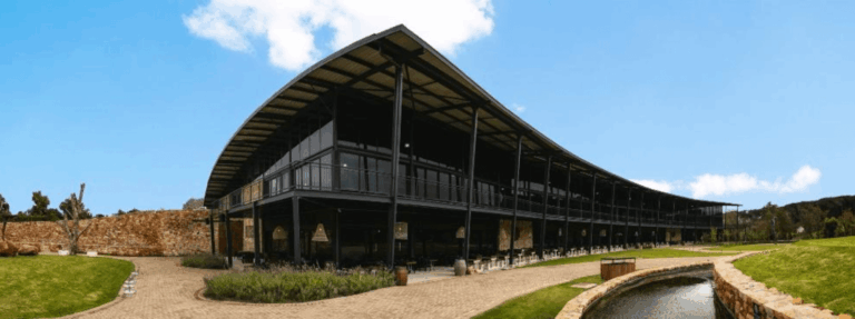 Modern black steel and glass winery building with a curved roof, surrounded by manicured lawns and stone paths. A narrow stream runs along the front, while a rustic stone wall borders the property under a bright blue sky.